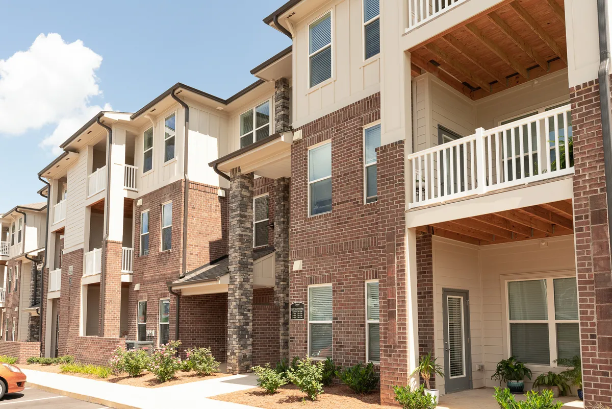 A welcoming view of the building’s brick facade with balconies overlooking green landscaping. Residents are seen relaxing on balconies and socializing in the courtyard, illustrating vibrant community life and the property’s appealing environment.