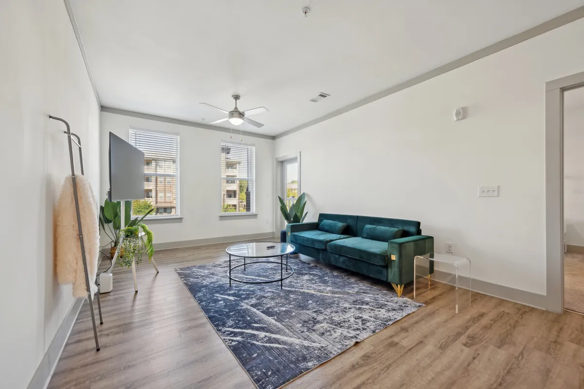 The image showcases a stylish living room with a plush teal sofa centered around a round coffee table, a wall-mounted TV perfect for movie nights, and vibrant plants by large windows that let in a warm sunset glow. The space feels both modern and cozy, inviting guests to relax or enjoy intimate gatherings.
