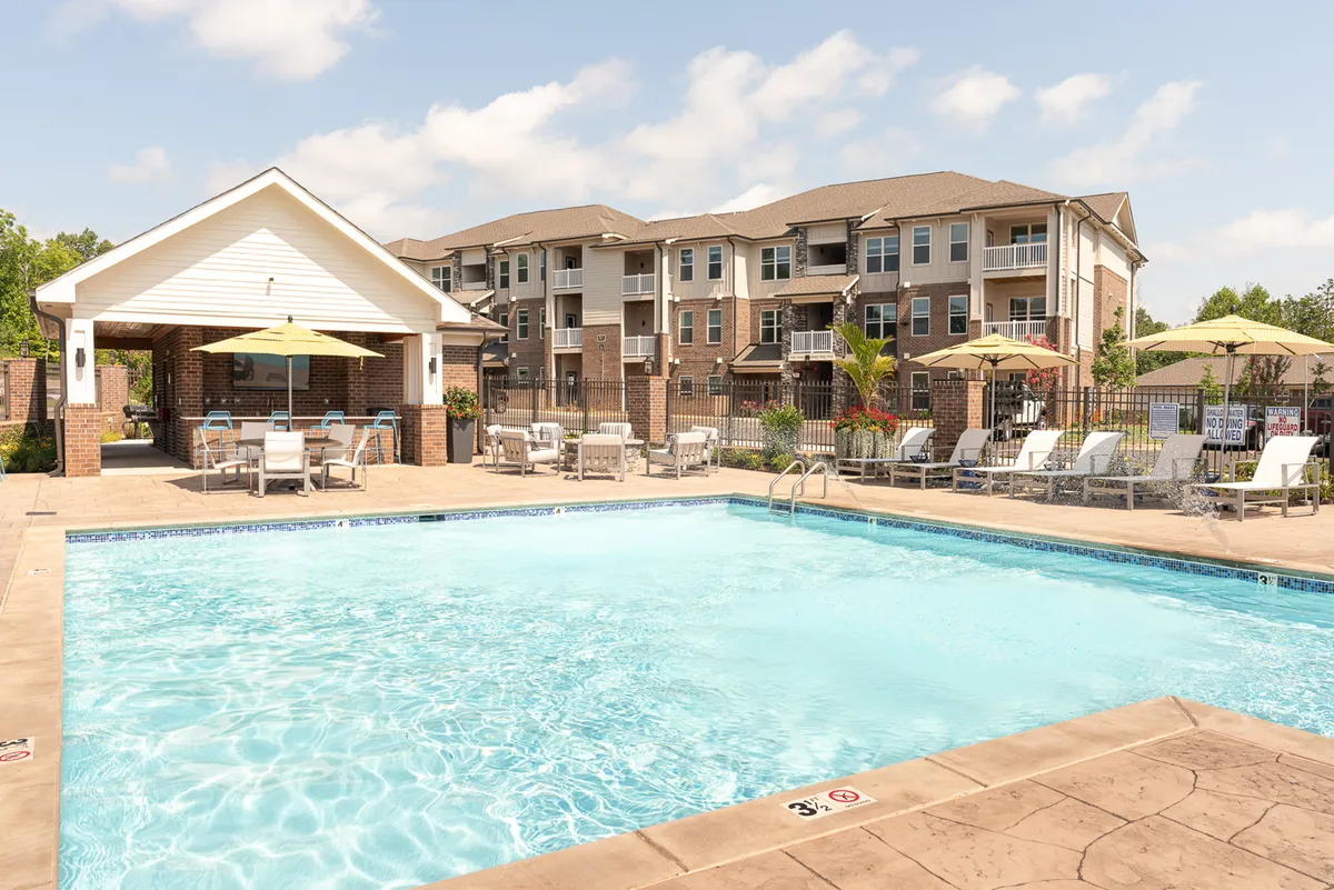 A sparkling community swimming pool surrounded by comfortable lounge chairs and a covered seating area. The pool is the centerpiece, with clear blue water reflecting the sky. The surrounding area features greenery and spaces for residents to relax or socialize, evoking a sense of leisure and community.
