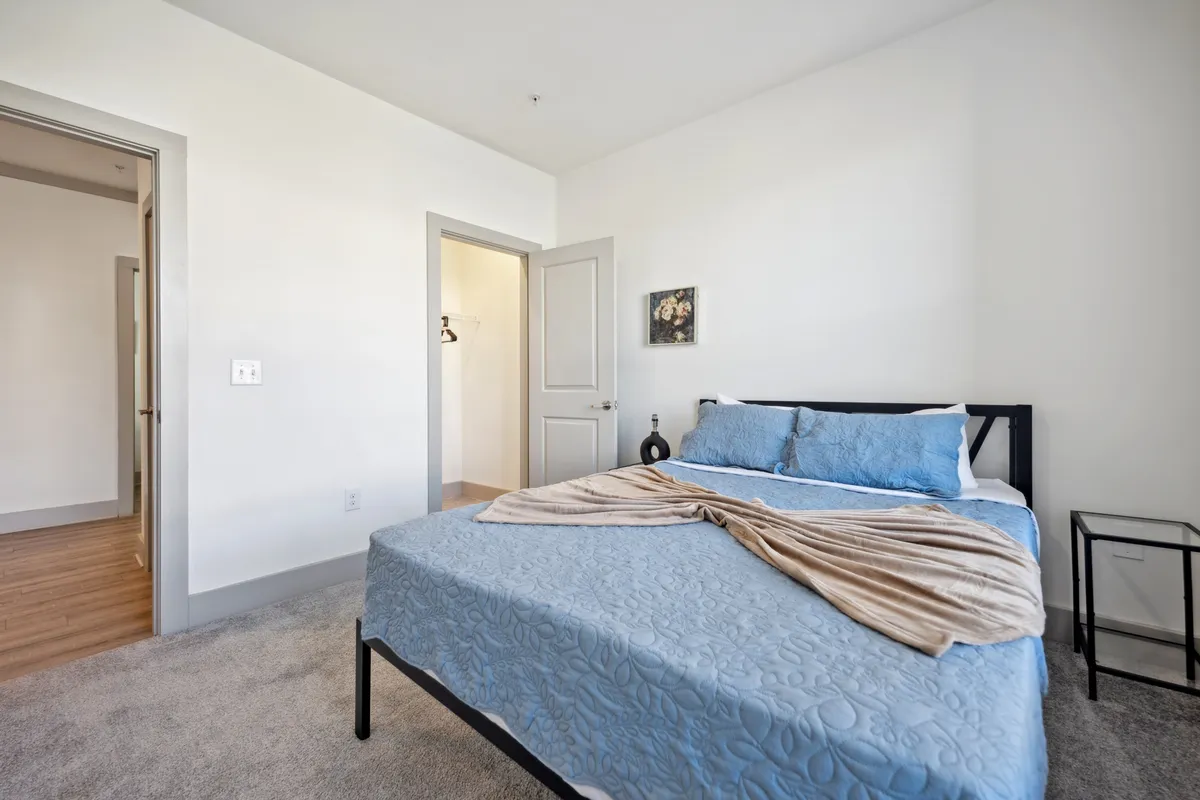 A cozy bedroom with blue bedding, decorative wall art, and a bedside table. The room is bathed in soft light, creating a calm and inviting atmosphere ideal for restful sleep or early morning routines.