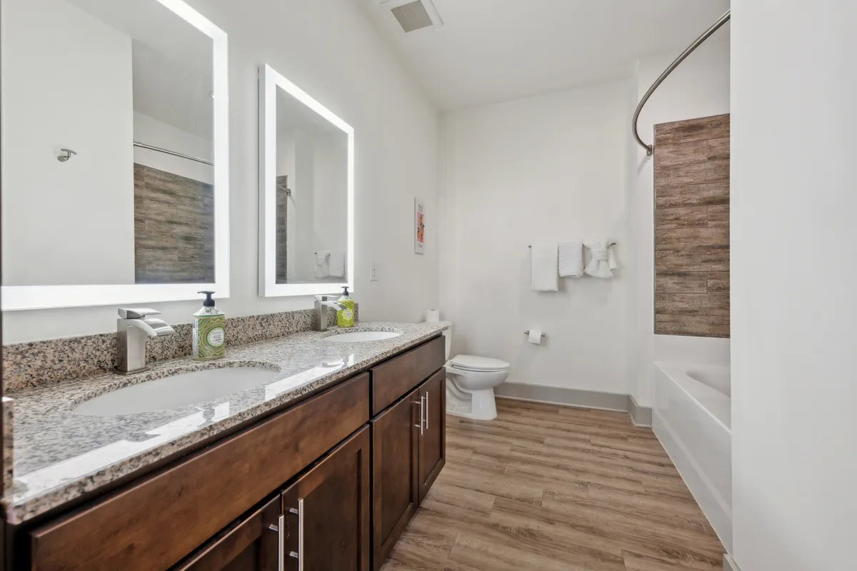 A spacious, sophisticated bathroom with a double vanity, granite countertop, large mirrors, wood-like flooring, and a luxurious bathtub. The space radiates calm and offers both functionality and indulgence.