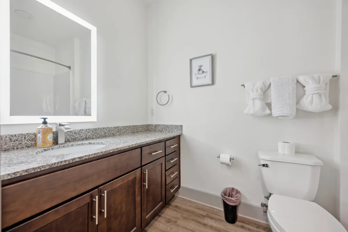 A contemporary bathroom scene featuring a granite countertop, illuminated mirror, framed artwork, and neatly arranged towels. The bathroom exudes freshness and is clearly designed for relaxation and self-care.