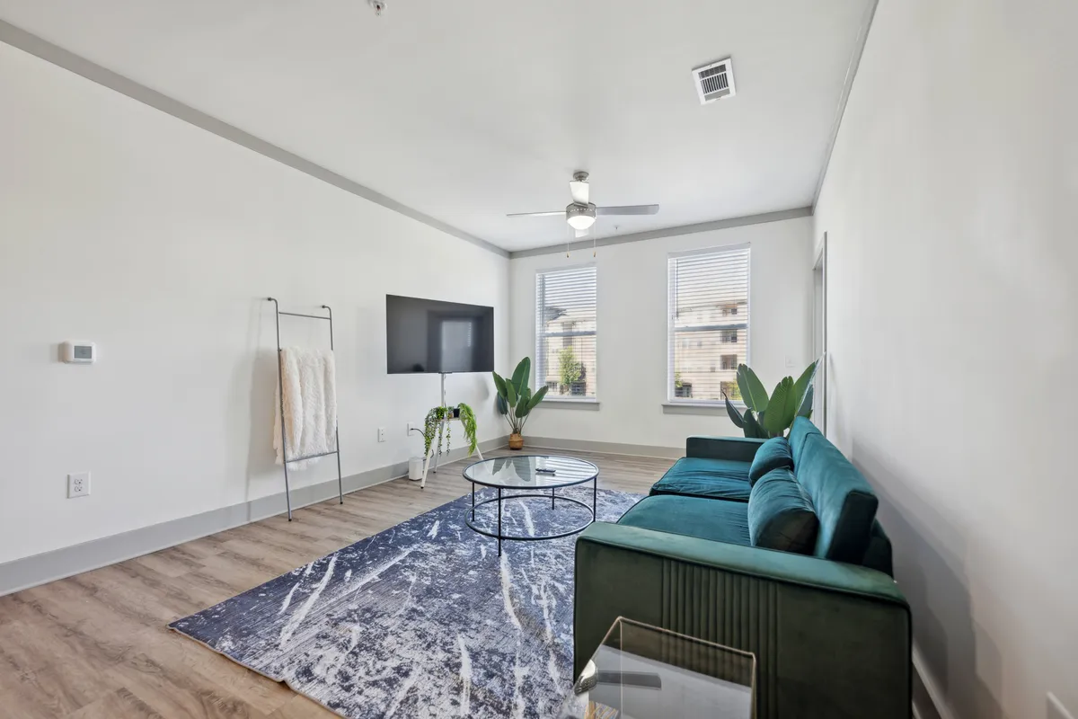 A tranquil living space featuring a teal sofa, glass coffee table, modern TV, decorative plants, and a soft area rug. The design blends style and comfort, illuminated by gentle daylight streaming in.