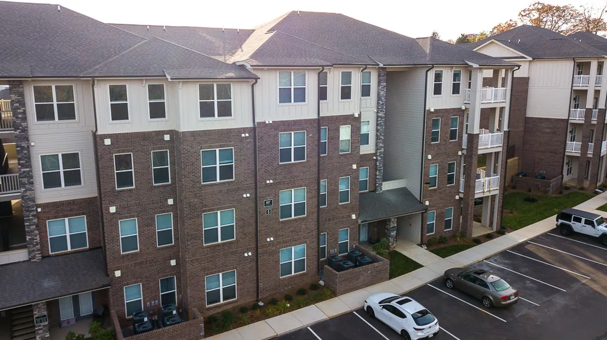A contemporary exterior shot showing modern architecture with large windows and a landscaped area. The image prominently features the parking lot, with cars parked conveniently close to the building, as well as inviting gathering spaces for residents.