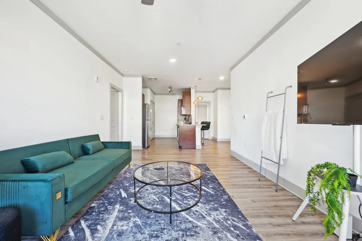 A modern living room featuring a plush green sofa, glass coffee table, area rug, and a wall-mounted television. The space is bathed in natural light from large windows, with contemporary décor and a cozy, inviting atmosphere.