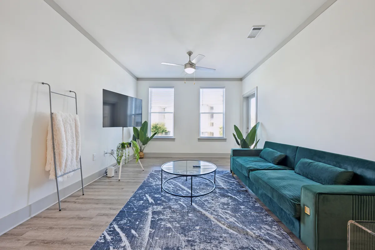 A stylish living room featuring a green sofa, a modern coffee table, large windows adorned with potted plants, and a wall-mounted TV. The space is bathed in natural light, creating an inviting and comfortable setting for relaxation or entertaining guests.