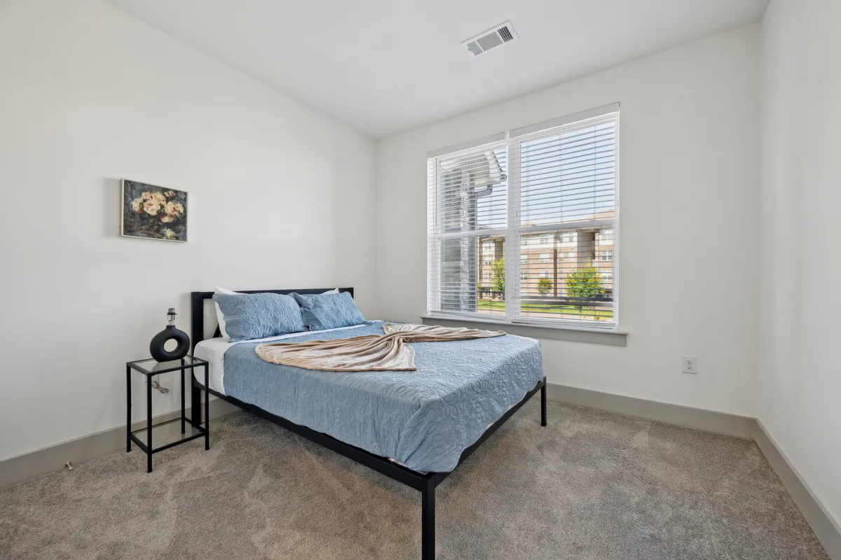 A tranquil bedroom featuring a bed with blue bedding, window with blinds allowing natural light, and tasteful artwork on the wall. The space feels peaceful and well-appointed, emphasizing comfort and style.